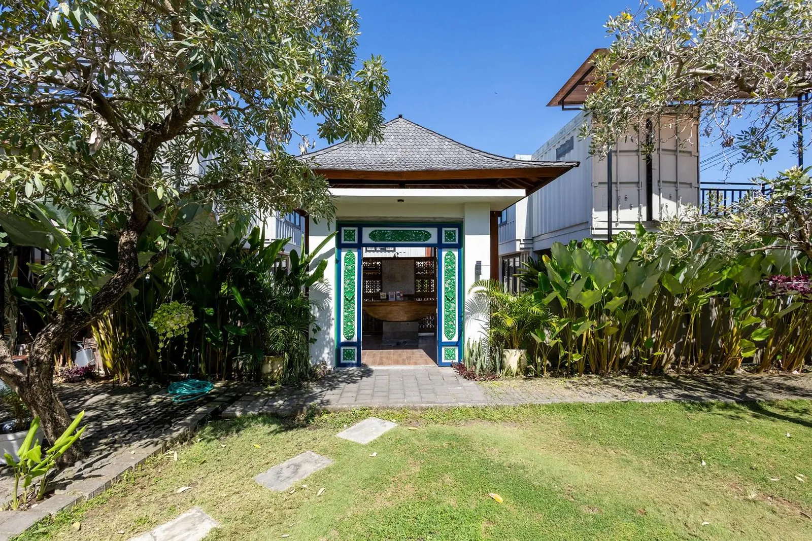 The garden pavilion with carved green doors, behind a lawn and frangipani trees
