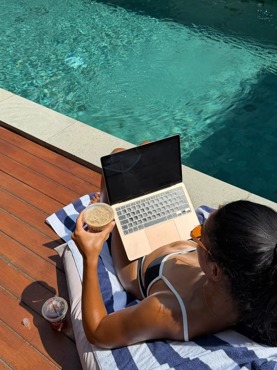 A guest working on a laptop on a striped towel beside the pool