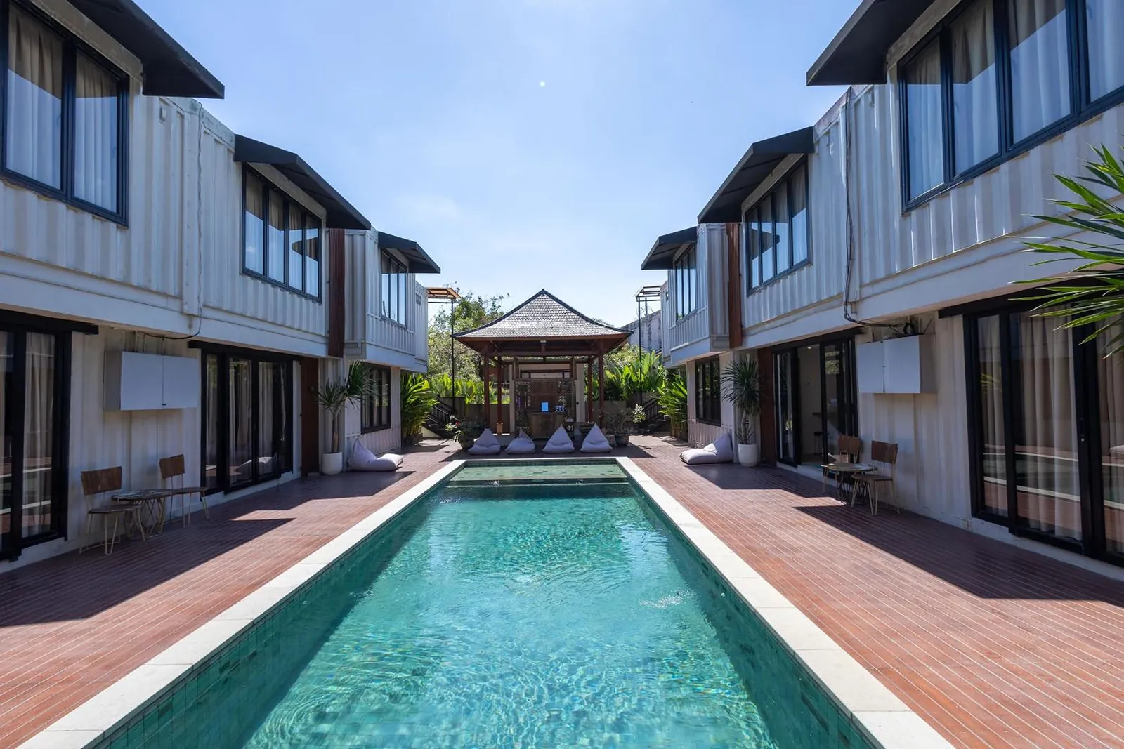 The long lap pool between two rows of white container suites, the thatched pavilion at its far end